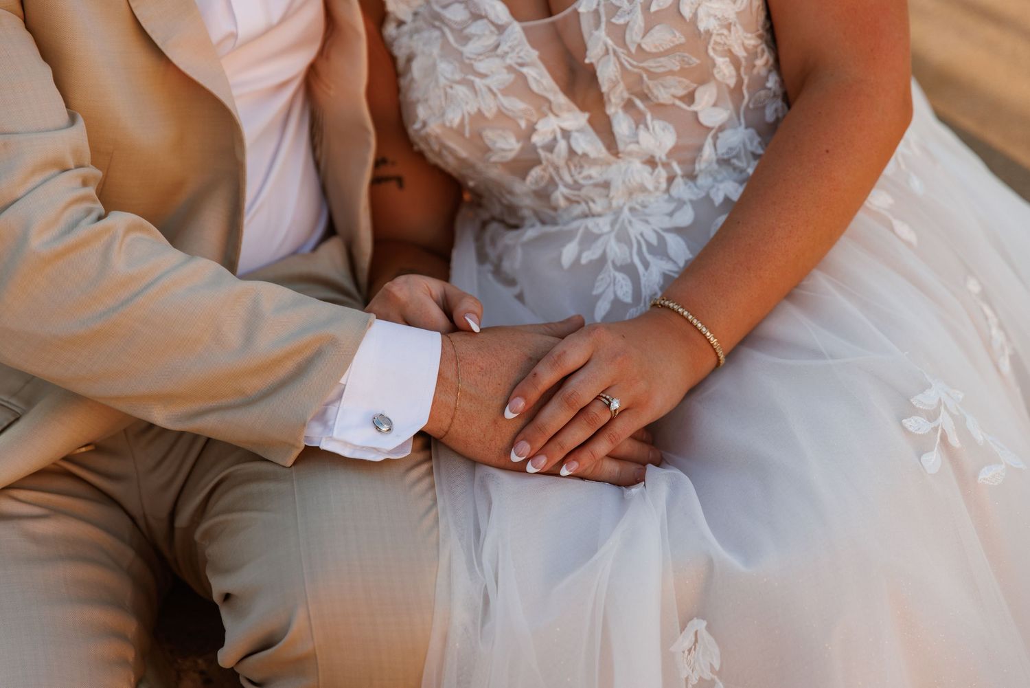 Close-up of bride and groom holding hands, showcasing wedding rings and elegant lace bridal gown.