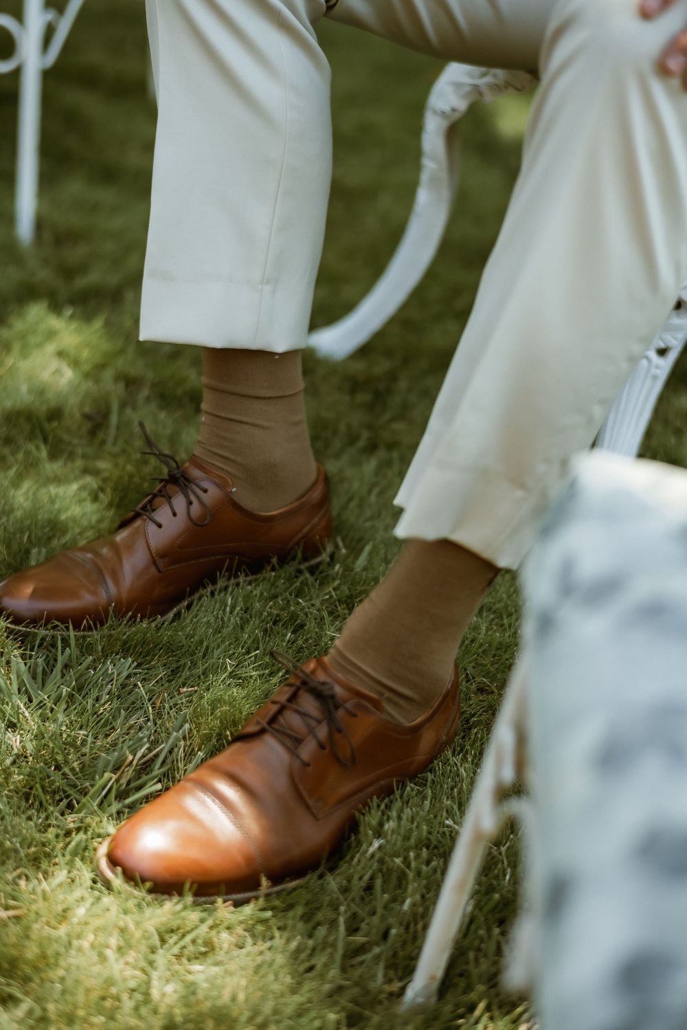 Close-up of brown leather lace-up dress shoes with tan socks and white trousers on green grass.