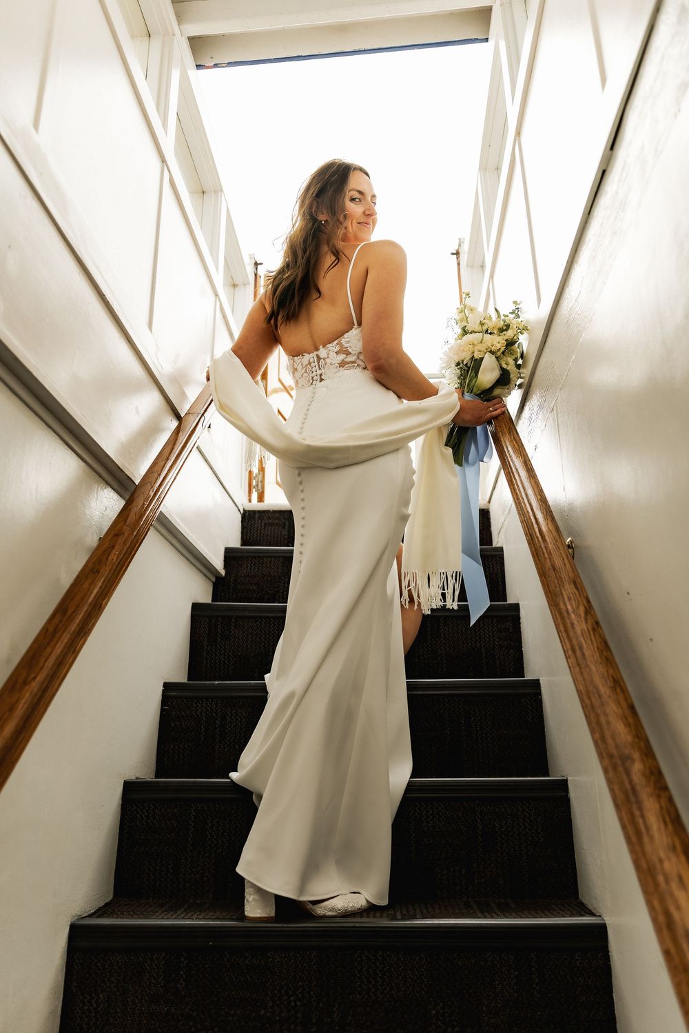 Bride in white gown holding bouquet on dark staircase, looking over shoulder with lace back detail and white wrap.