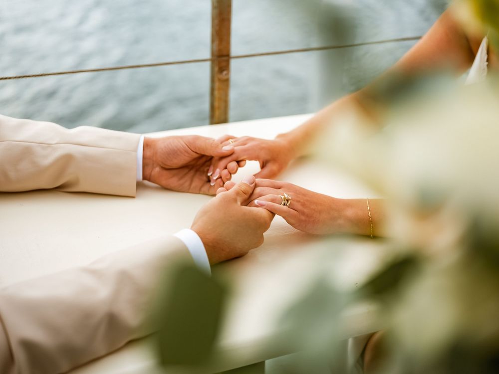 Bride and groom exchanging rings during outdoor wedding ceremony near waterfront, surrounded by greenery.