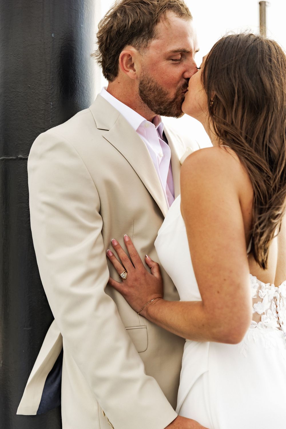Bride and groom sharing a romantic kiss, wearing white dress and beige suit, with wedding ring visible.