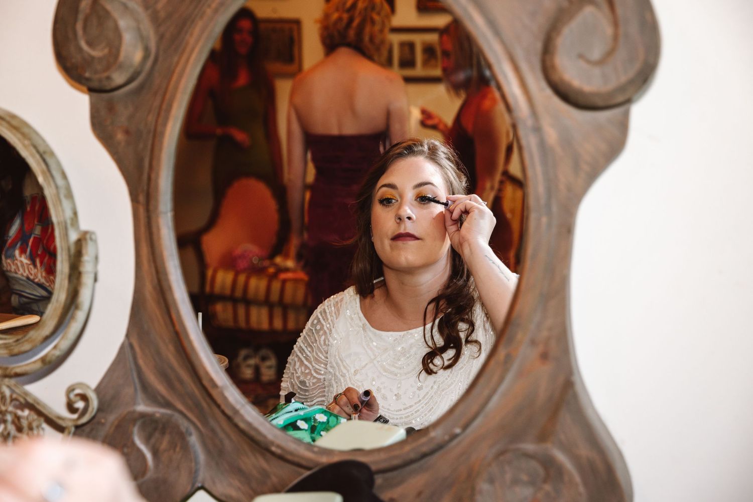 Bride applying makeup while looking into an ornate vintage mirror before her wedding.