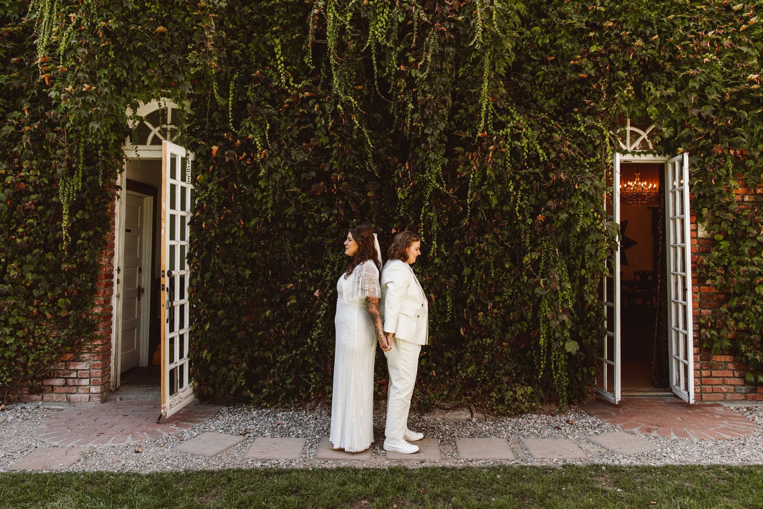 Two brides in white wedding attire standing back-to-back before a lush ivy-covered wall with French doors.