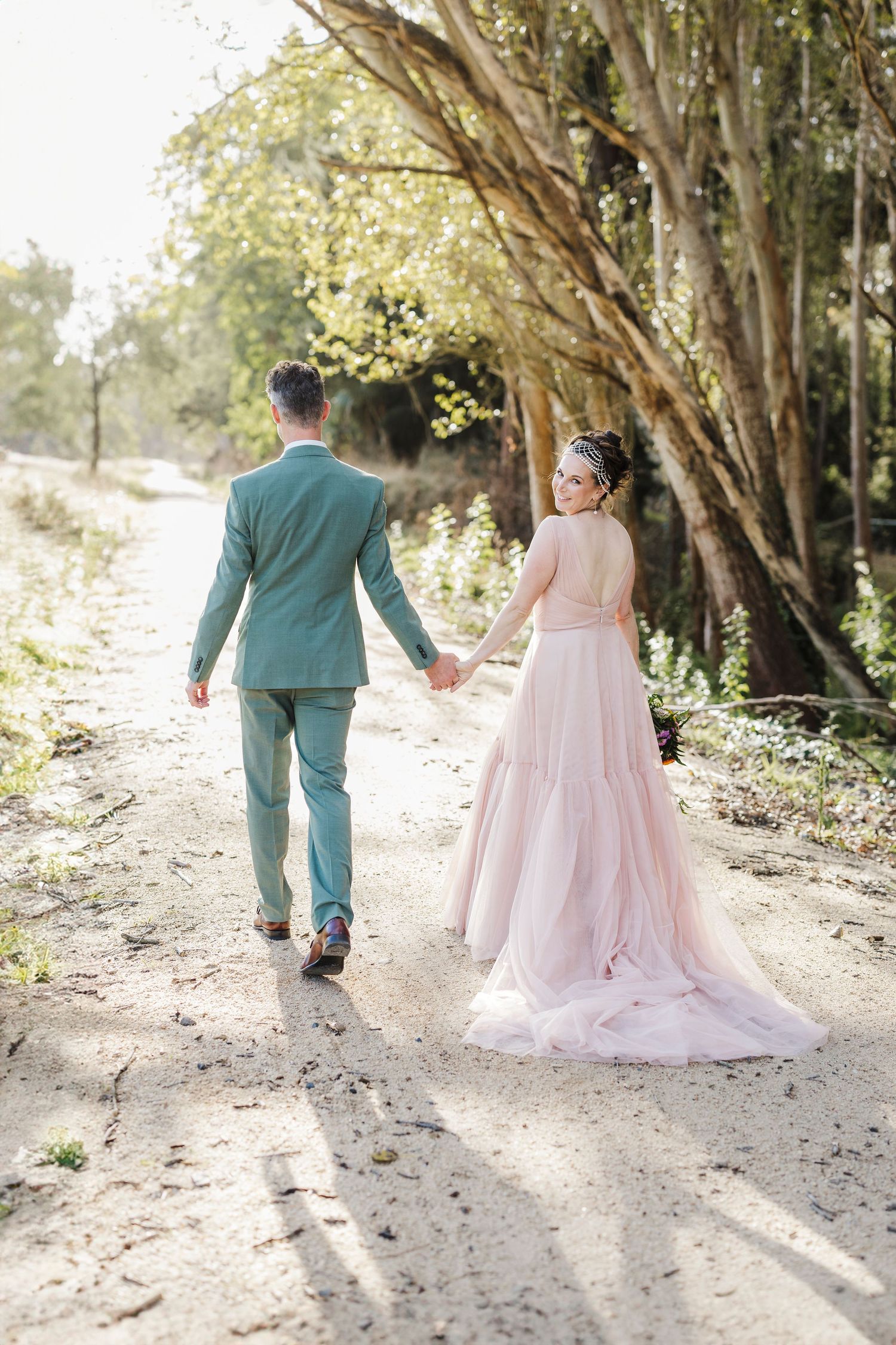 A couple in wedding attire walks hand in hand down a tree-lined path in soft sunlight.