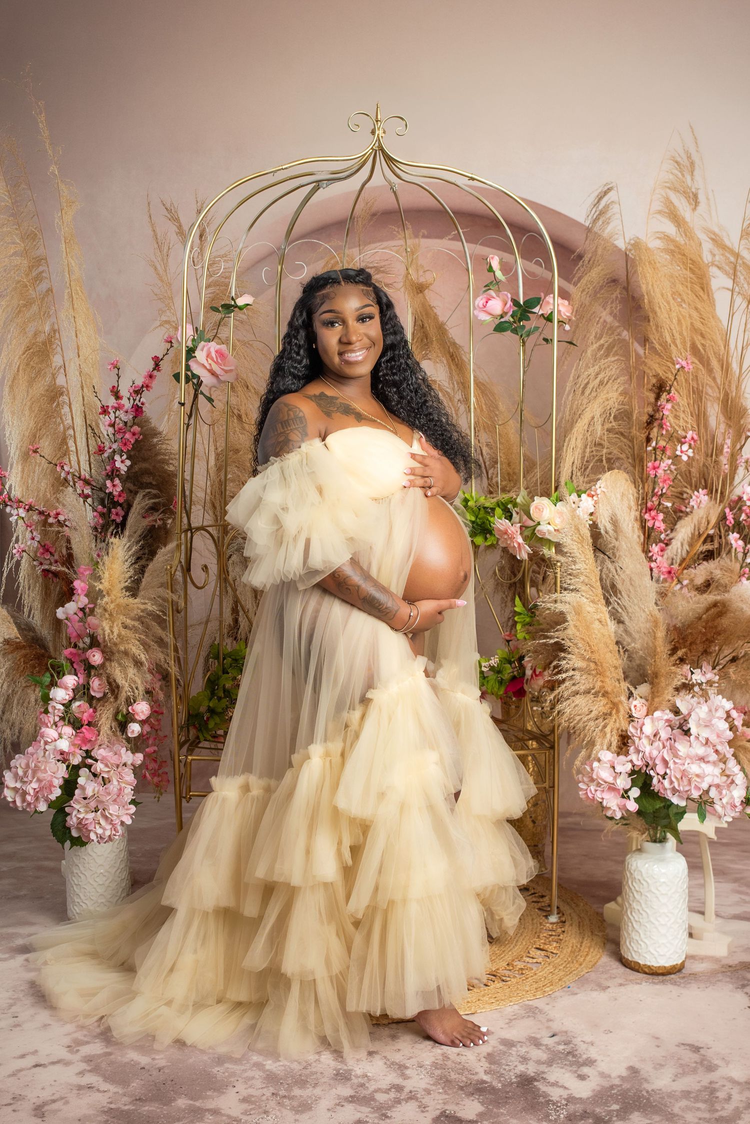 Pregnant woman in elegant cream tulle gown poses in floral studio setting with pampas grass and pink blooms.