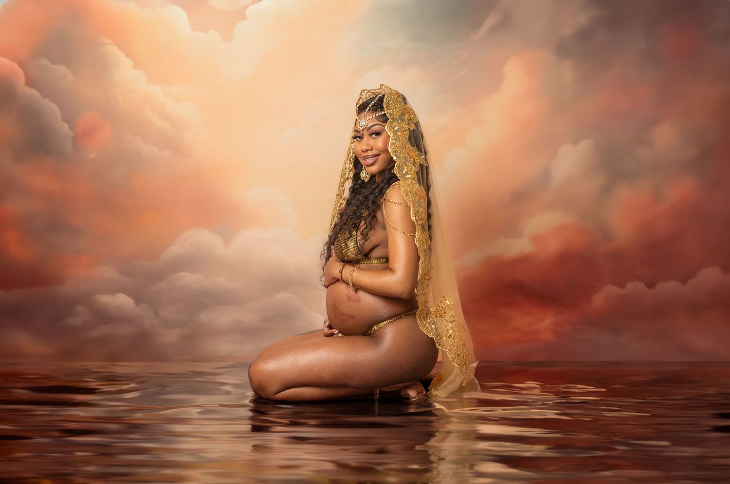 Artistic maternity photo of an expectant mother posing in water against dramatic orange and red cloudy sky at sunset.