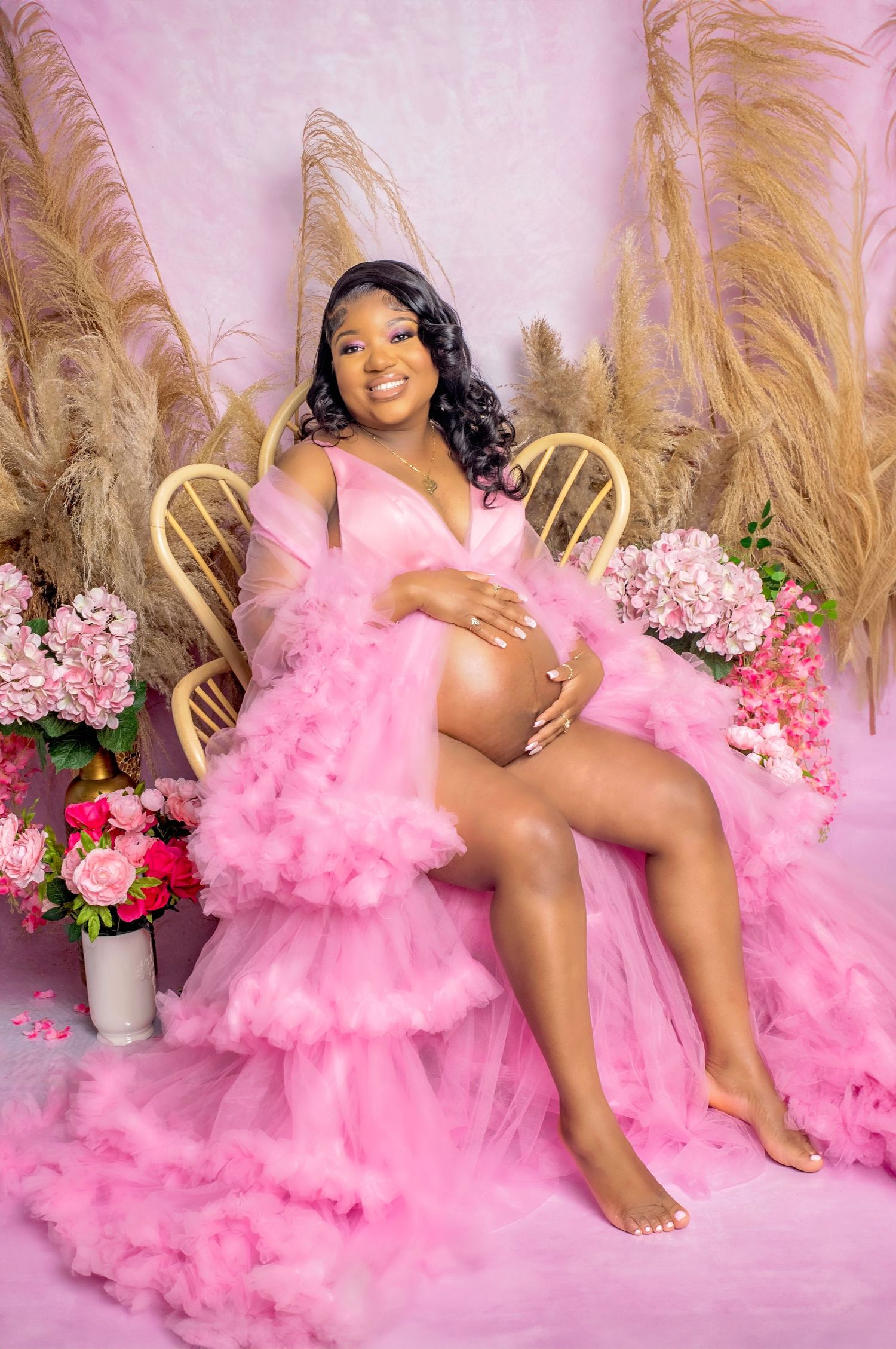 A maternity photoshoot with pink tulle dress against a floral backdrop with pampas grass decor and soft lighting.