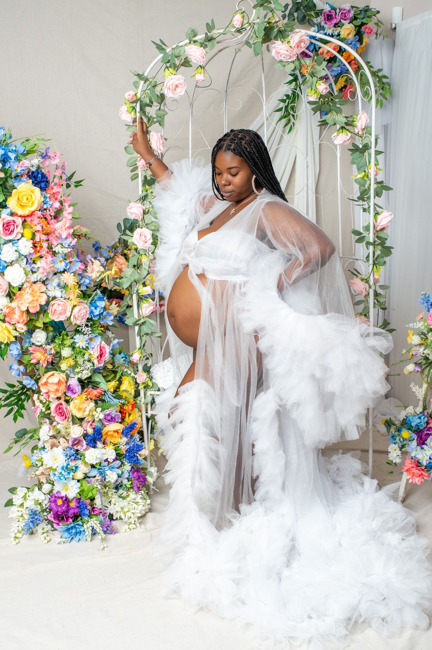 Ethereal maternity portrait with flowy white sheer gown against floral arch backdrop with colorful spring blooms.