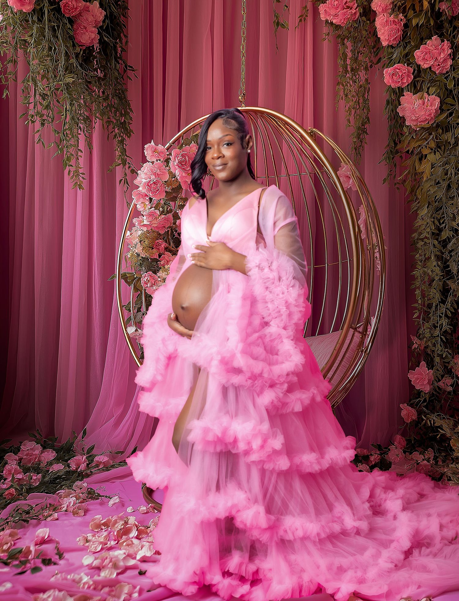 Pregnant woman in pink ruffle gown sitting in gold circular swing chair surrounded by pink flowers and draping.