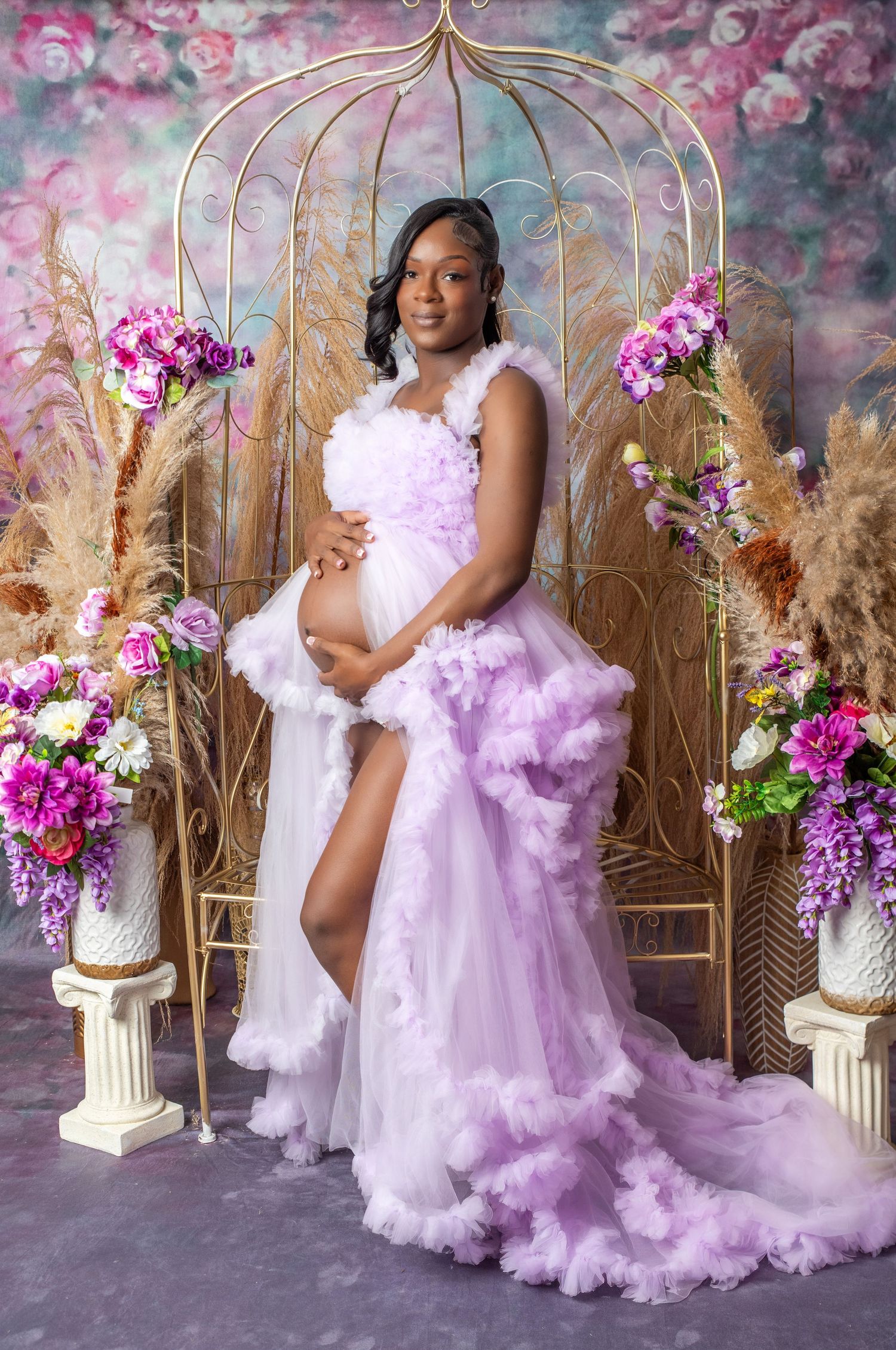 Pregnant woman in lavender ruffle gown poses regally among pink flowers and gold arch for maternity photo.