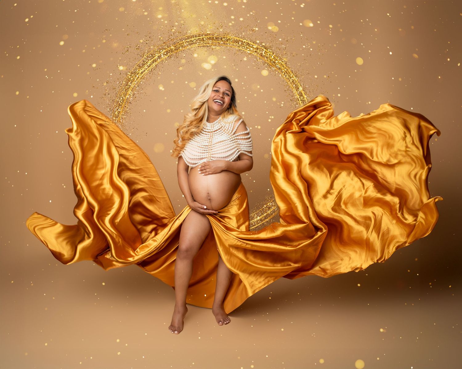 Artistic maternity photo with flowing golden fabric and glittering lights against warm brown background.