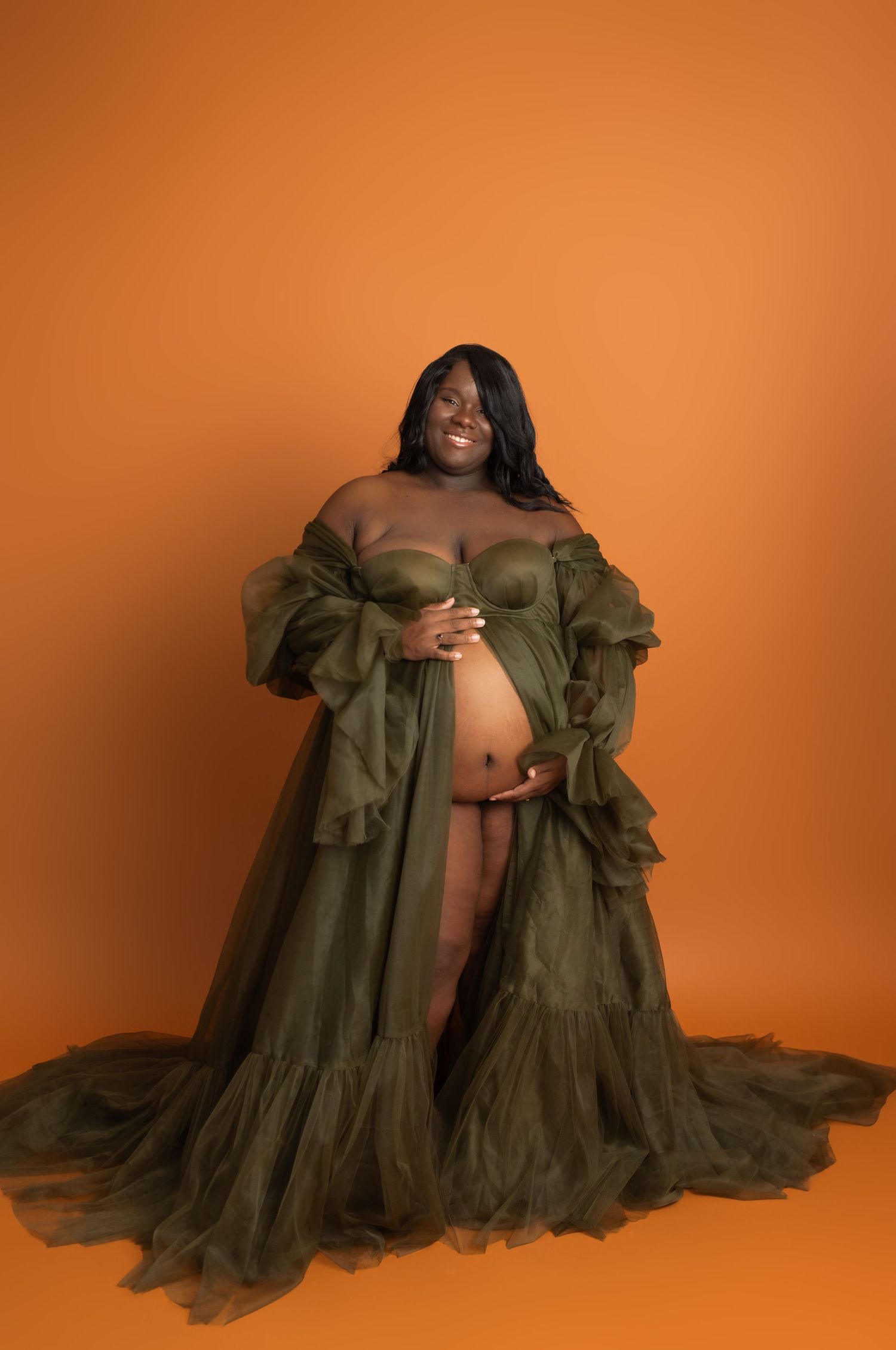 Pregnant woman in olive green tulle maternity gown poses against orange backdrop in professional studio photo.