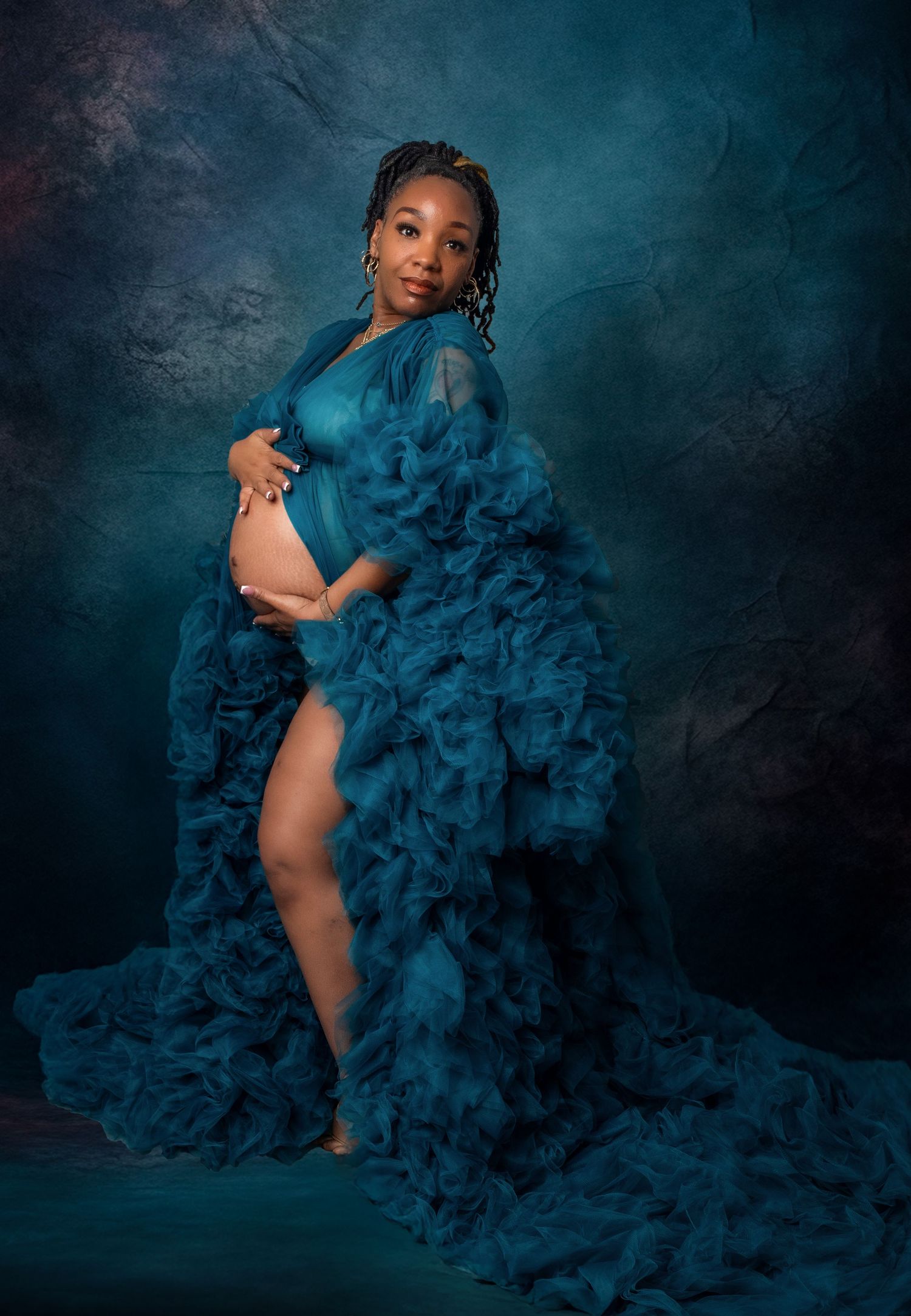 Dramatic maternity photoshoot in teal featuring flowing tulle fabric against a dark moody backdrop.