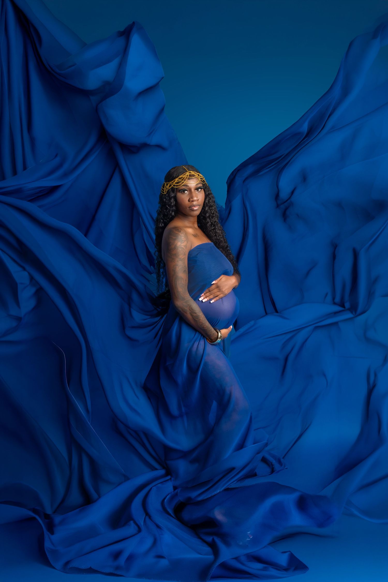 Pregnant woman in flowing royal blue fabric gown with gold headpiece poses gracefully against blue backdrop.