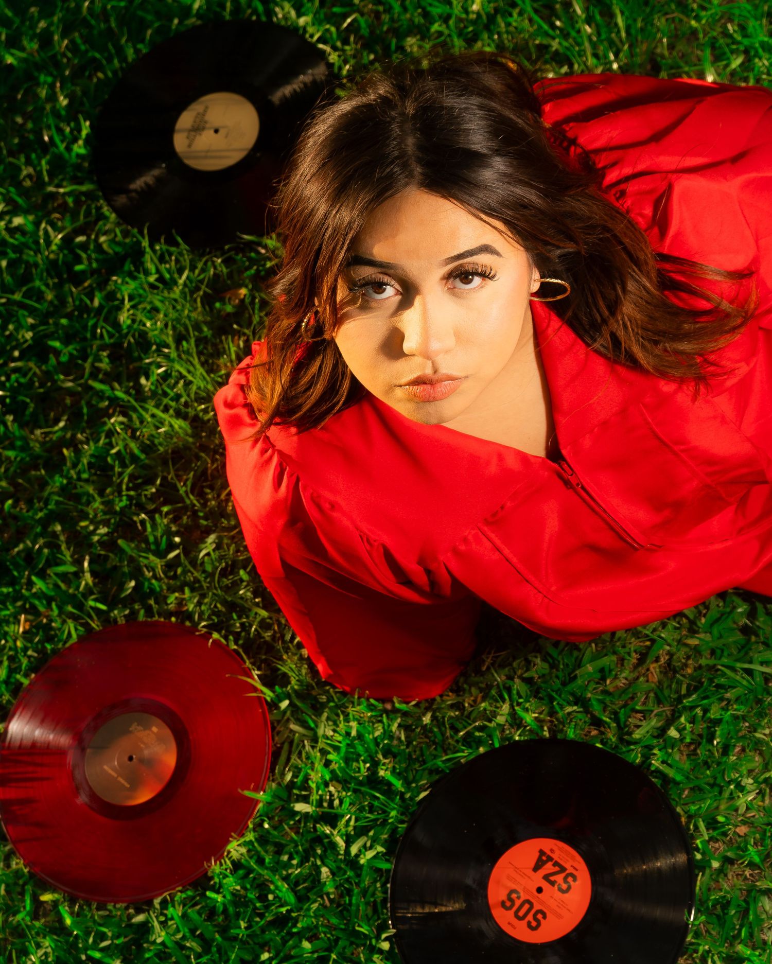 Young woman in red jacket lying on grass surrounded by vinyl records, looking up at camera with a pensive expression.