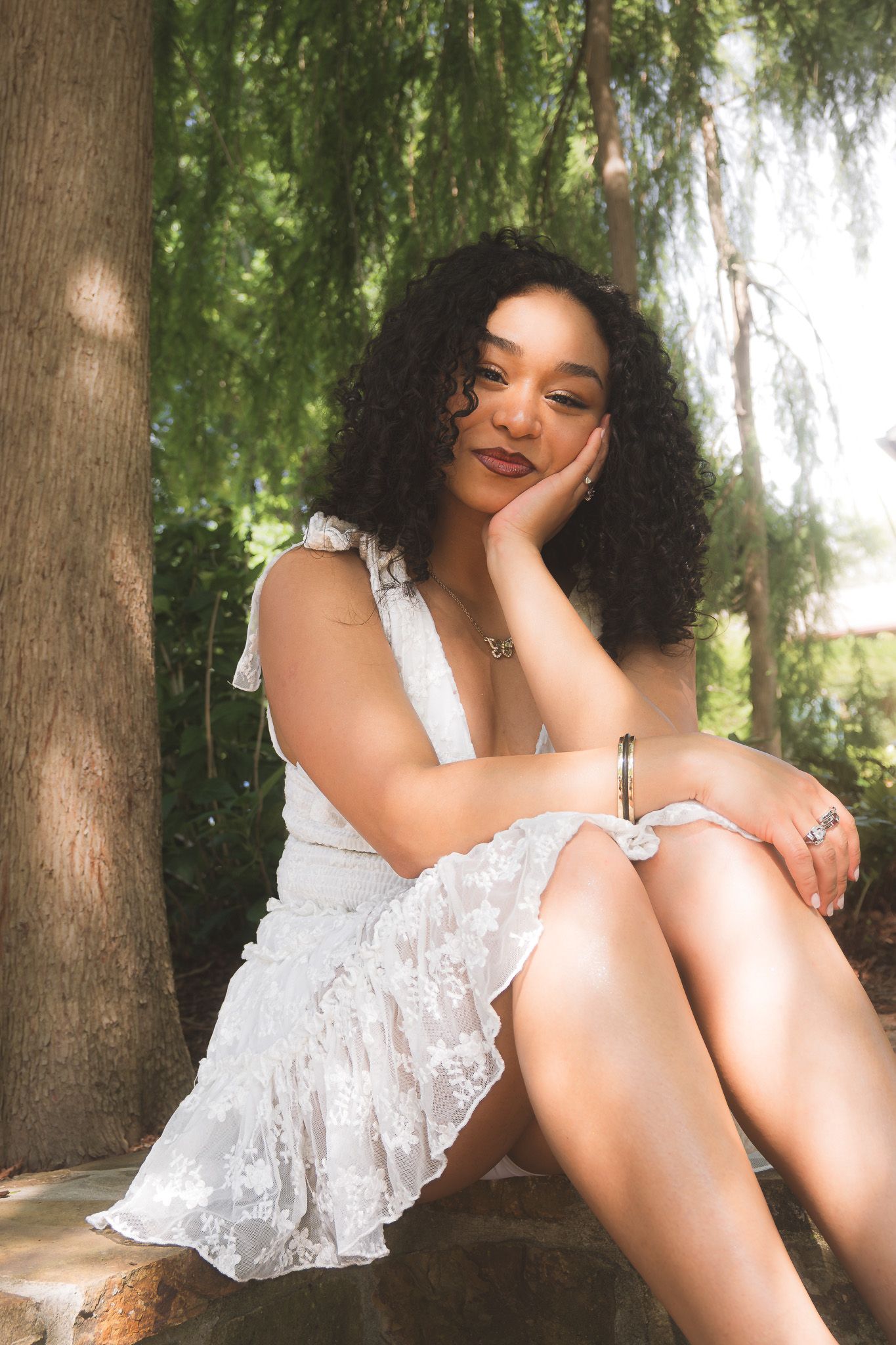 A person in a white lace dress sits on the ground beneath a willow tree in a dreamy outdoor photoshoot.