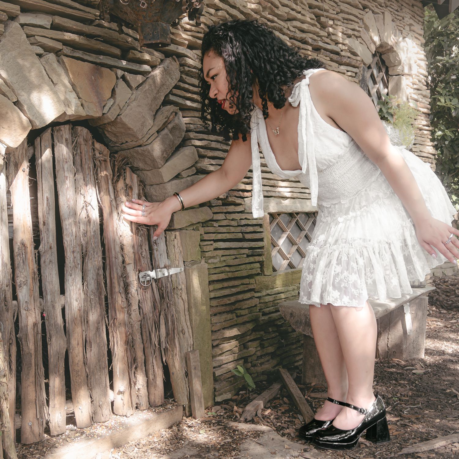 A person in a white lace dress bends down to examine an old wooden fence in a rustic outdoor setting.