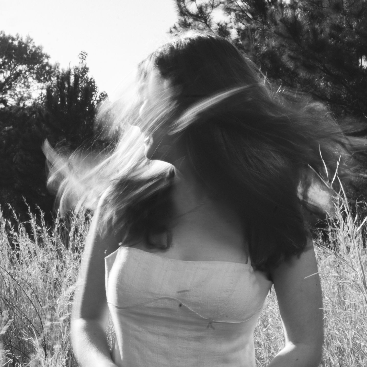 Black and white photo of a woman with long hair blowing in the wind, standing in a field with trees behind her.