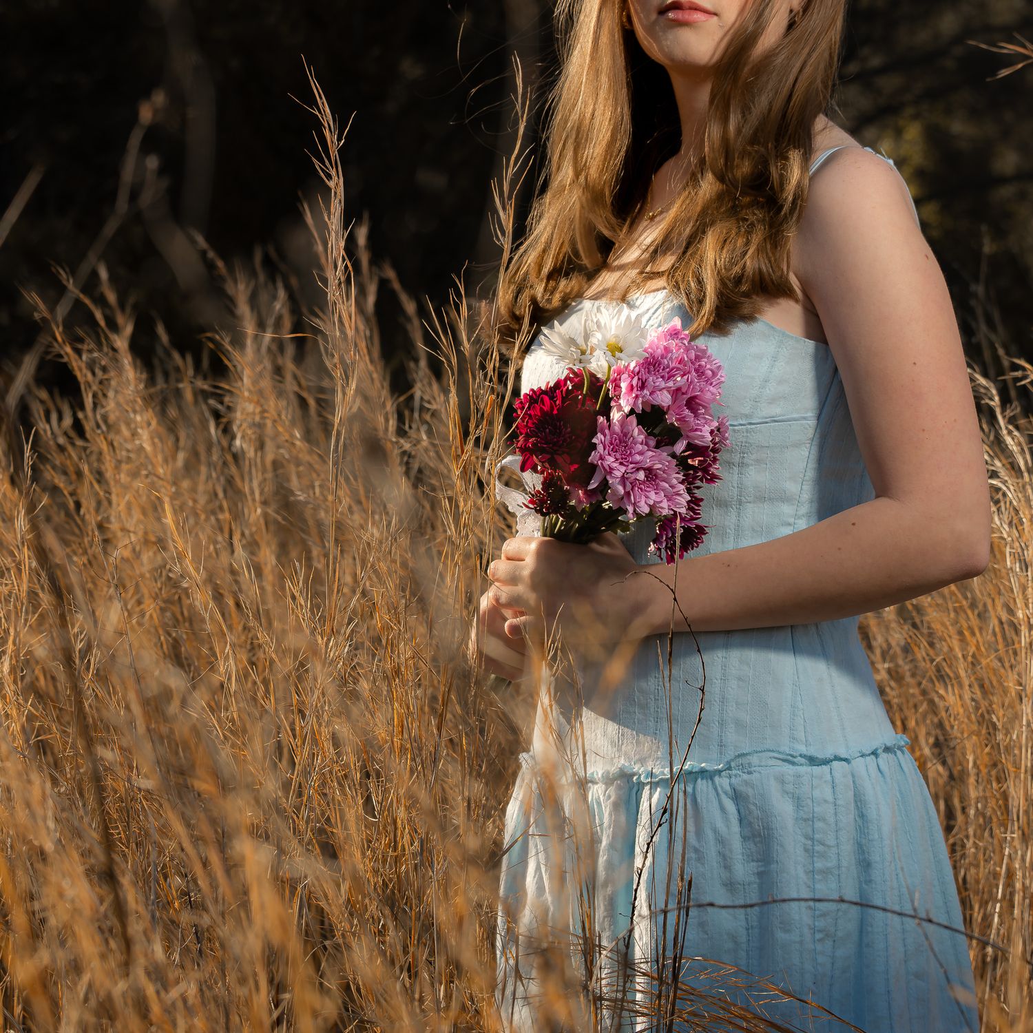 Young woman in light blue dress holding pink and red flower bouquet standing in golden dry grass field.