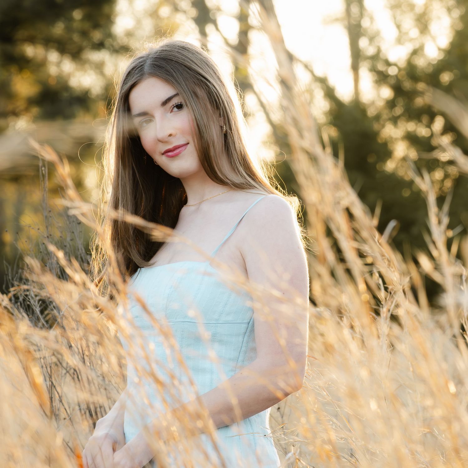 Portrait in a light dress standing among golden tall grass at sunset with soft natural lighting.