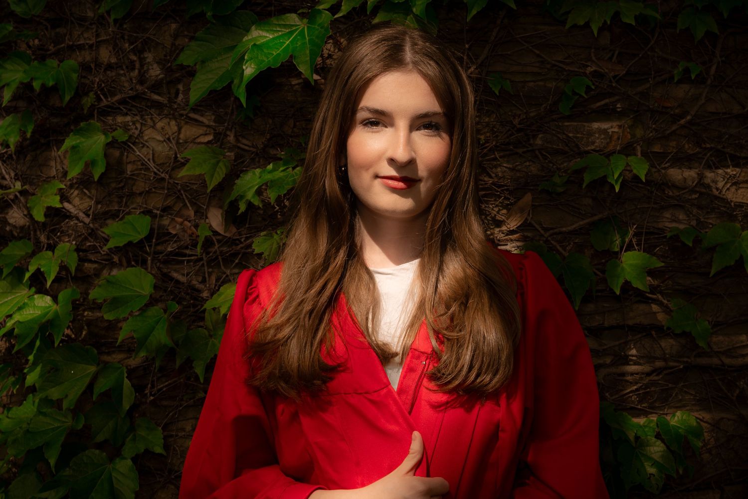 Young woman with long brown hair wearing a red jacket, posing outdoors against a lush green ivy-covered wall.