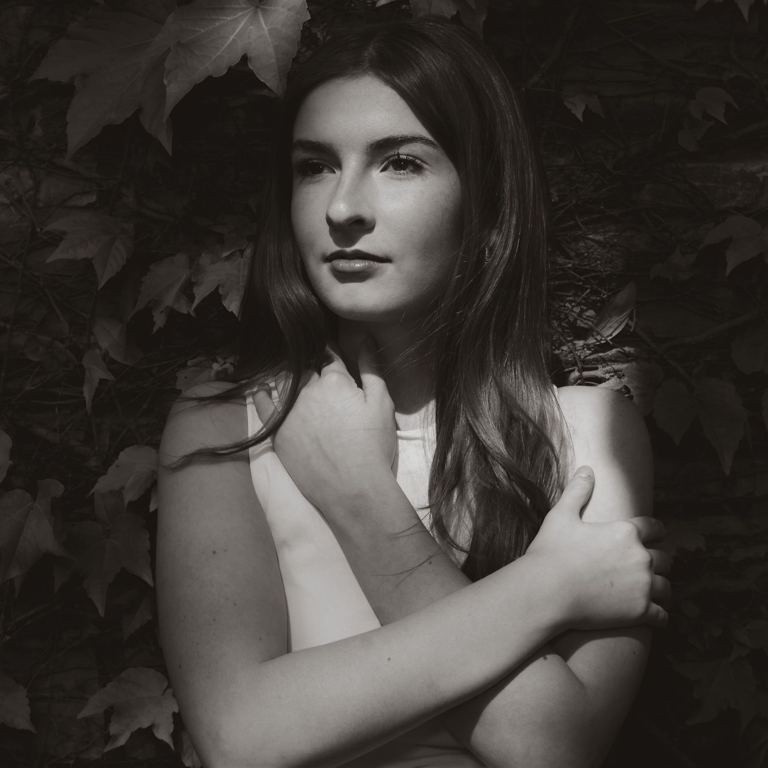 Black and white portrait of a young woman with long hair, arms crossed, surrounded by lush ivy leaves.