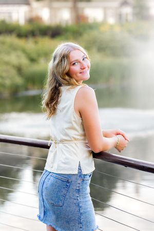 Woman in white sleeveless top and denim shorts poses joyfully by a lake during an outdoor photoshoot.