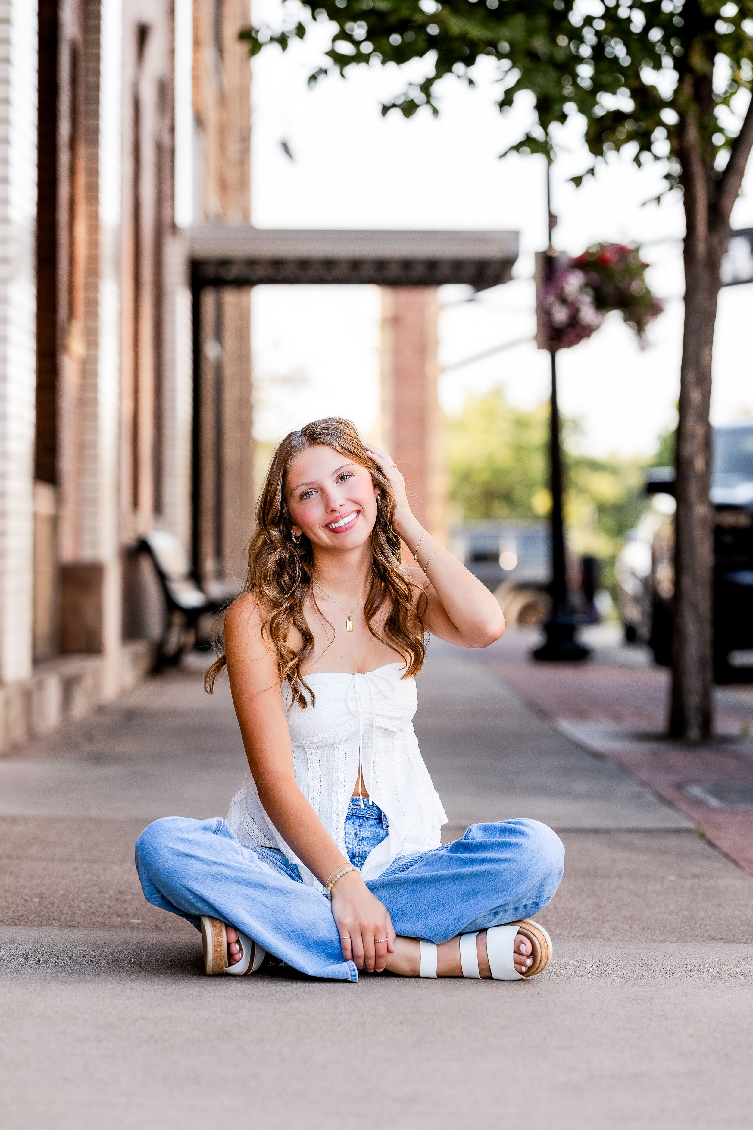 A person in white top and blue jeans sits casually on a sidewalk with columns and trees in an urban setting.
