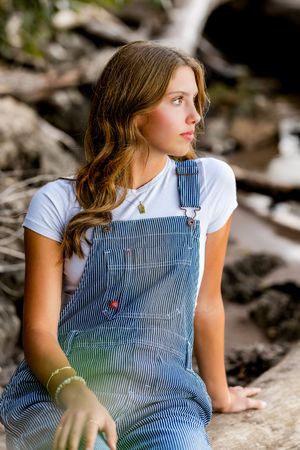 Someone wearing blue and white striped overalls and a white t-shirt sits thoughtfully on rocks in a natural setting.