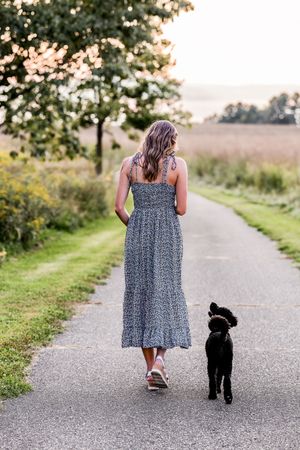 A person in a floral dress walks down a country path with a black dog on a summer evening.