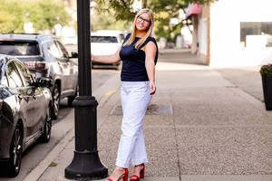 Person wearing white pants and black top walks down urban sidewalk with parked cars in background.