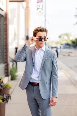 A well-dressed person in a light gray suit and sunglasses poses confidently on a city sidewalk.