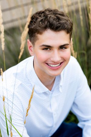 Young person in white shirt smiles warmly while sitting outdoors with natural grasses in background.