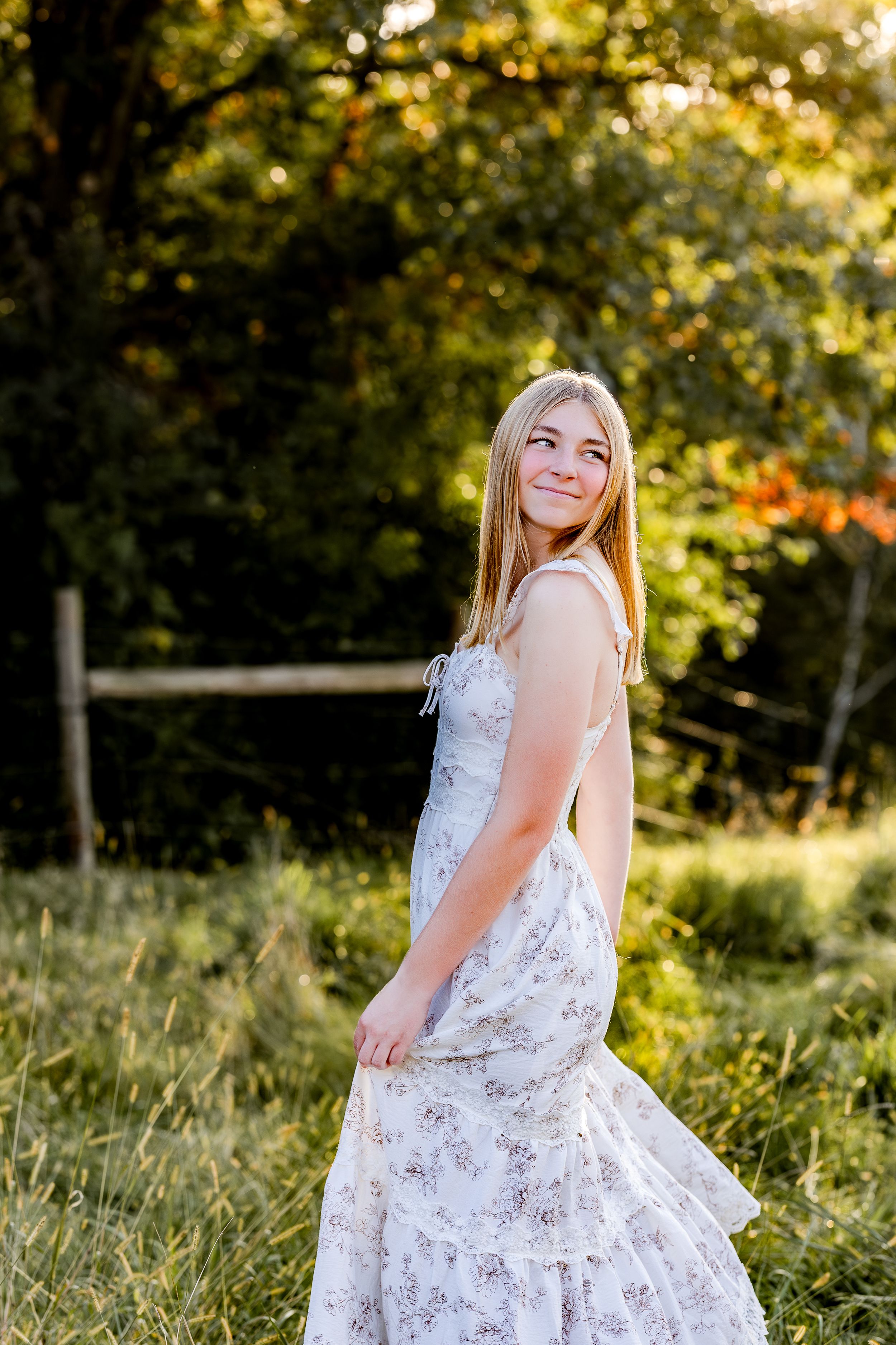 A person in a white floral dress poses in a sunlit meadow during golden hour.