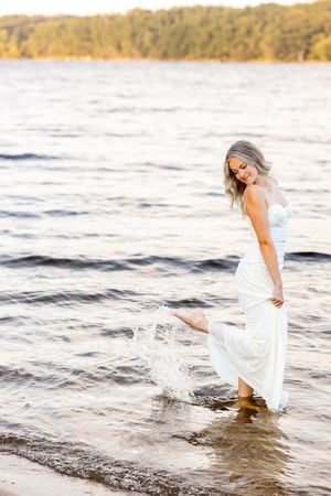 A person in a white dress splashes playfully in the water at sunset along a scenic shoreline.