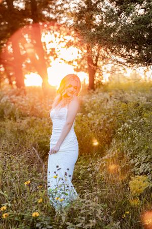 A person in a white dress stands among wildflowers and tall grass during a golden sunset in a dreamy outdoor setting.