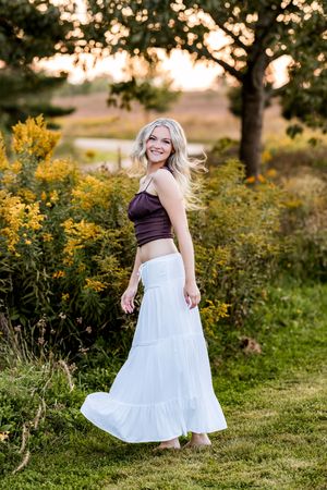 A person in a brown top and white flowing skirt twirls in a field of yellow wildflowers at sunset.