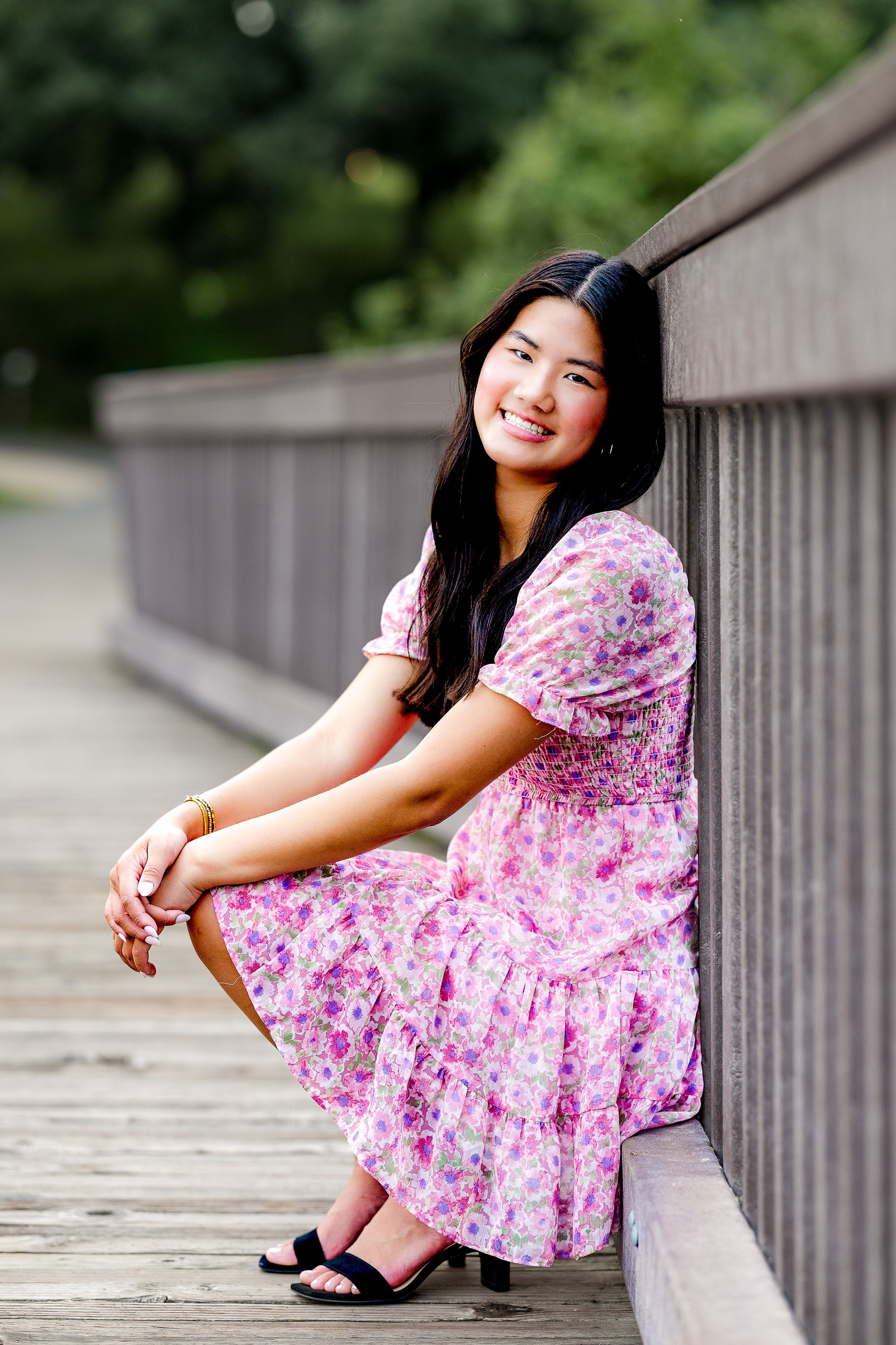 A person in a pink floral dress sits on a wooden bridge railing while smiling on a summer day.