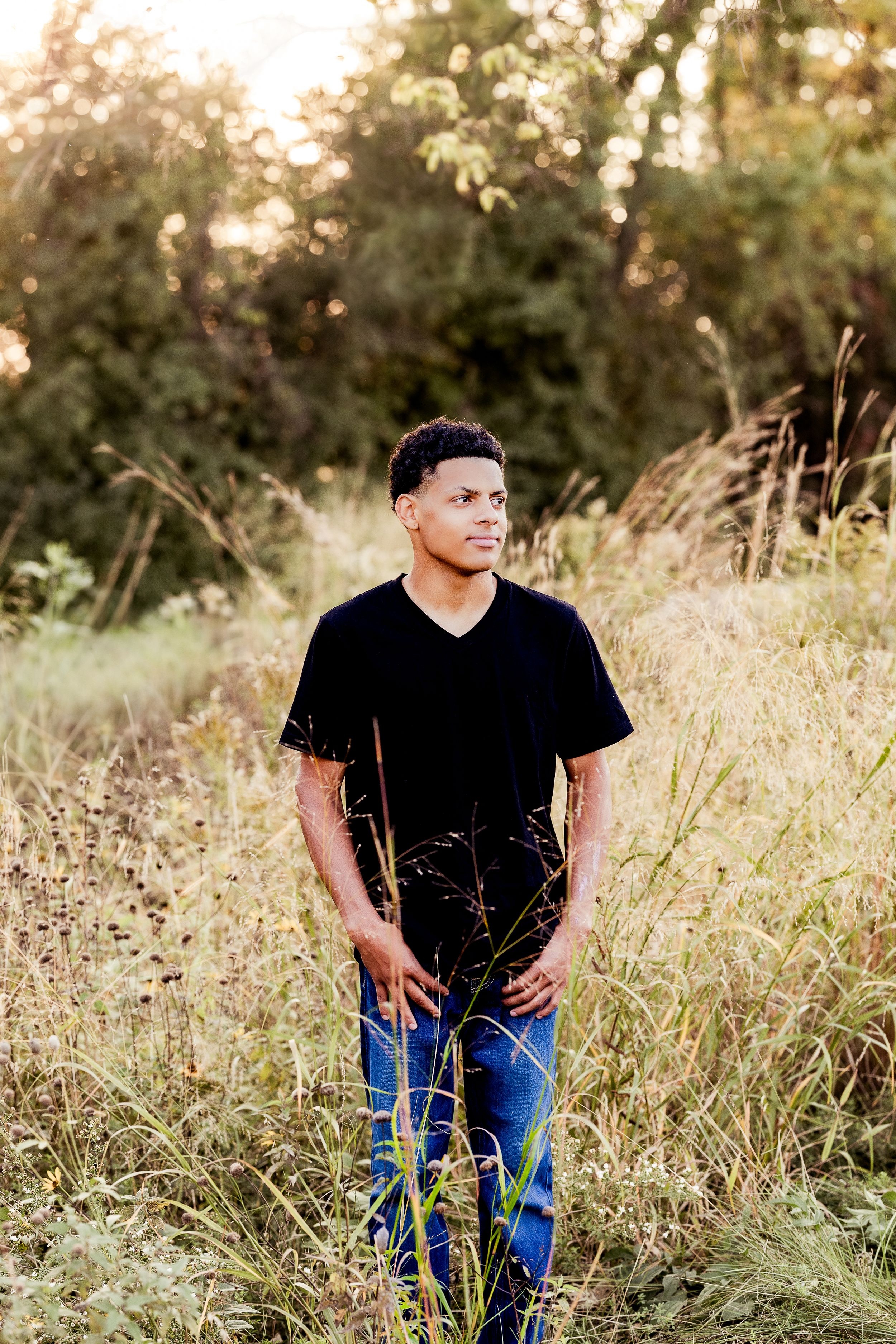 Person in black shirt and jeans poses in tall grass during golden hour sunset in outdoor portrait session.