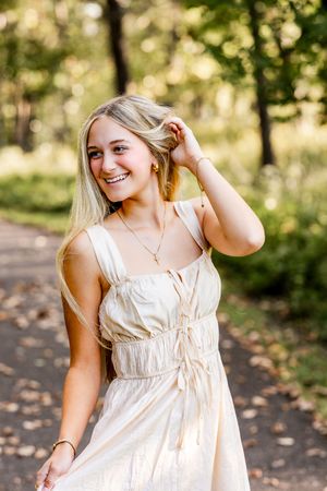 A person in a flowing cream-colored summer dress poses outdoors on a wooded path with dappled sunlight.