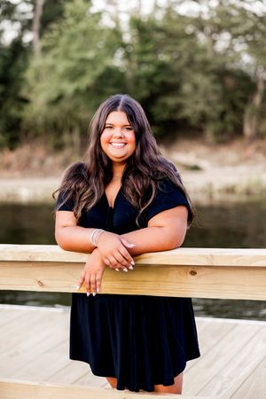 A person in a navy blue dress smiles while standing on a wooden deck overlooking a lake.