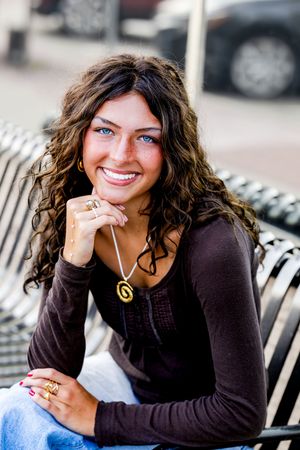 A person in a brown sweater sits outdoors smiling with curly hair and a necklace in a casual portrait series.