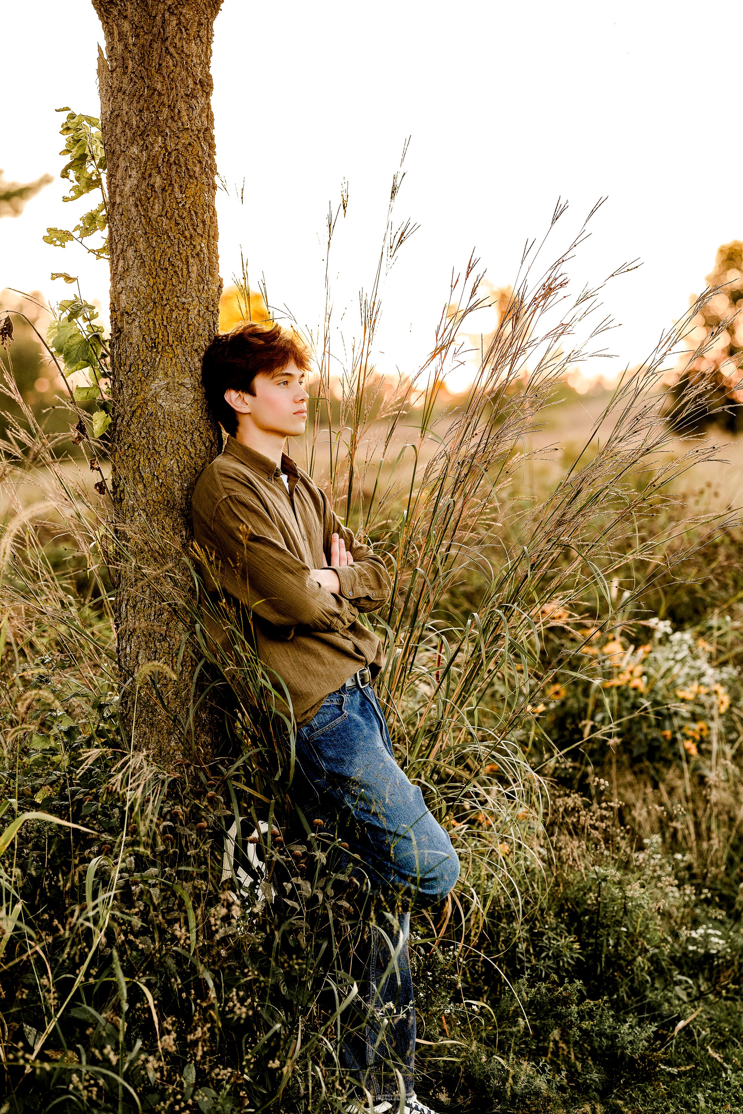Person in brown jacket and jeans sits thoughtfully against tree trunk in tall grass at sunset.