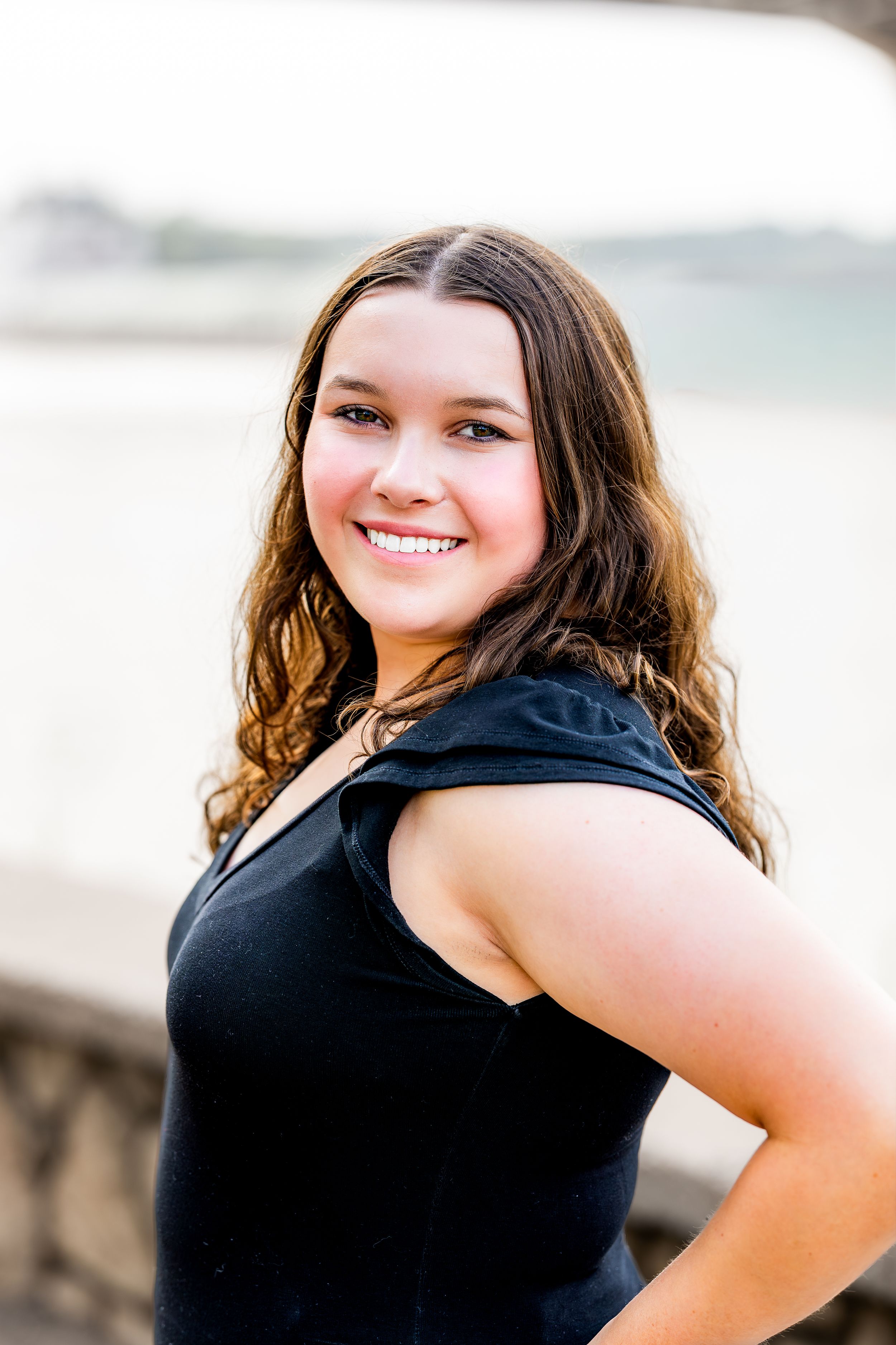 Portrait of someone with long curly brown hair wearing a black sleeveless top posing outdoors with a bright smile.