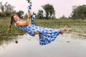 A person in a blue floral dress swings playfully over a pond while holding onto a rope swing on a summer day.