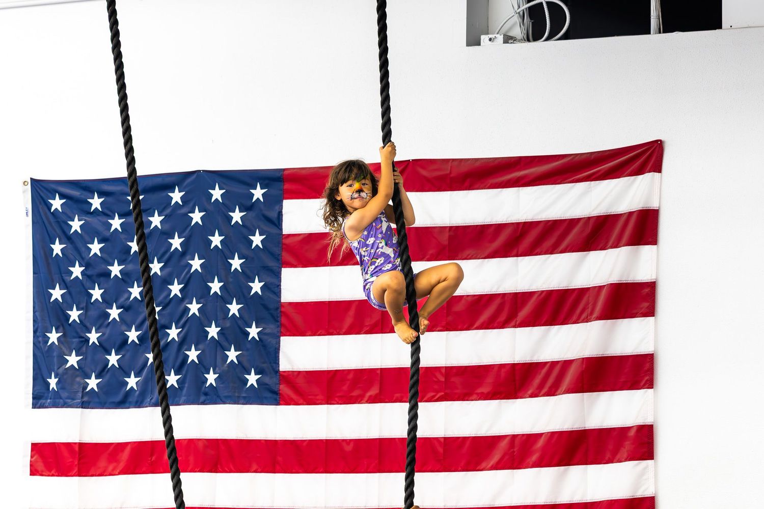 San Diego sports photography of a young girl climbing a rope during a gymnastics class with a large American flag in the background.