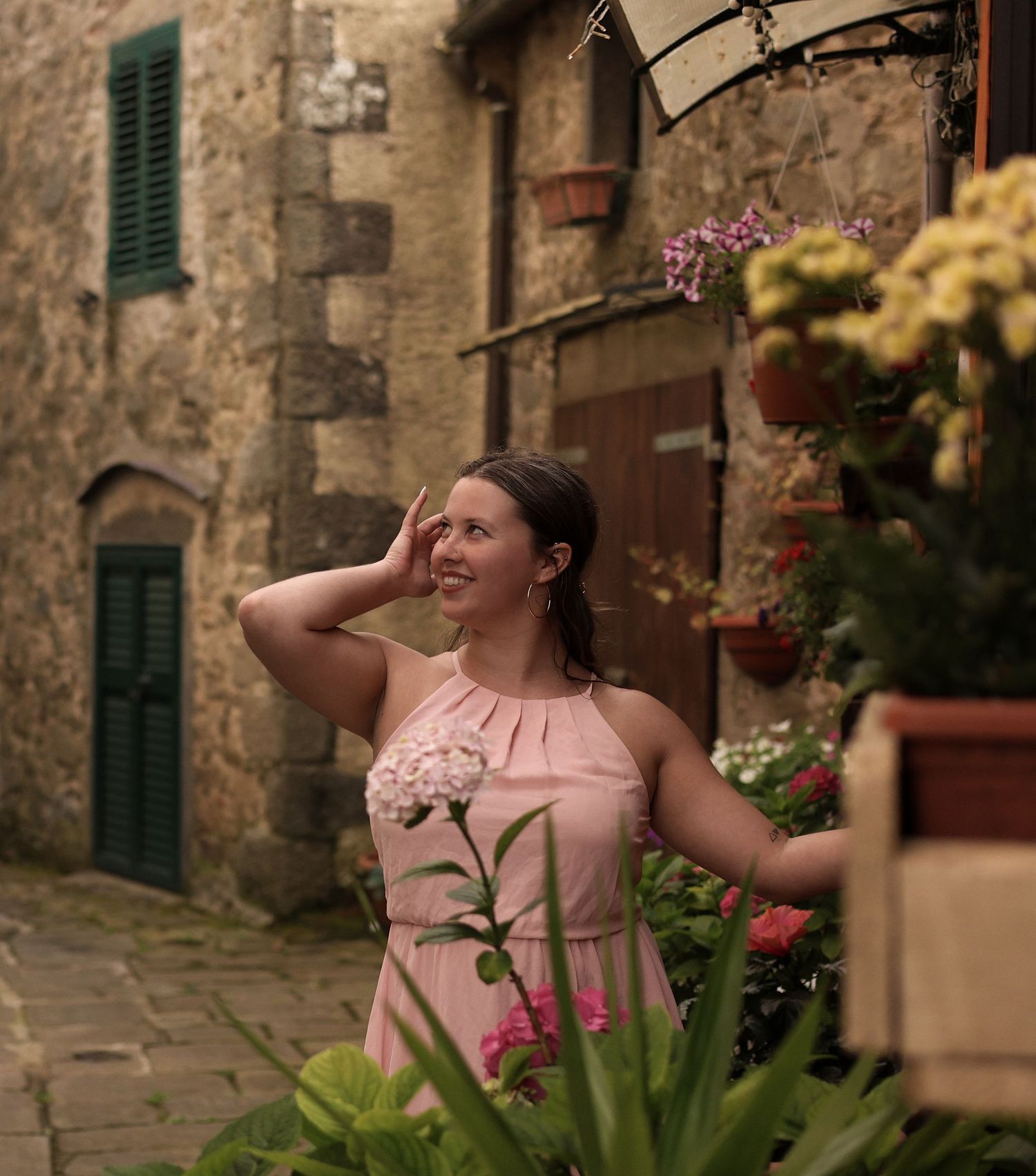 A person in a pink dress poses playfully against a rustic stone wall with green shutters and potted flowers.