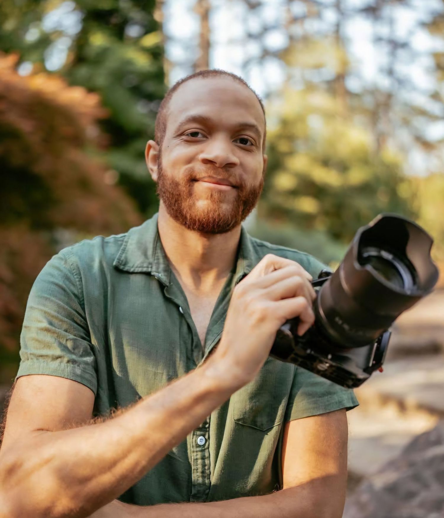 A photographer with a ginger beard smiles while holding a professional camera outdoors in natural lighting.