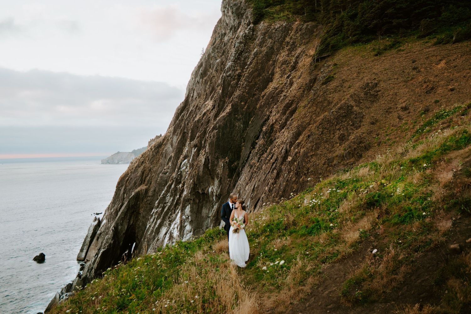 Couple in wedding attire standing on a dramatic coastal cliff path overlooking the ocean.