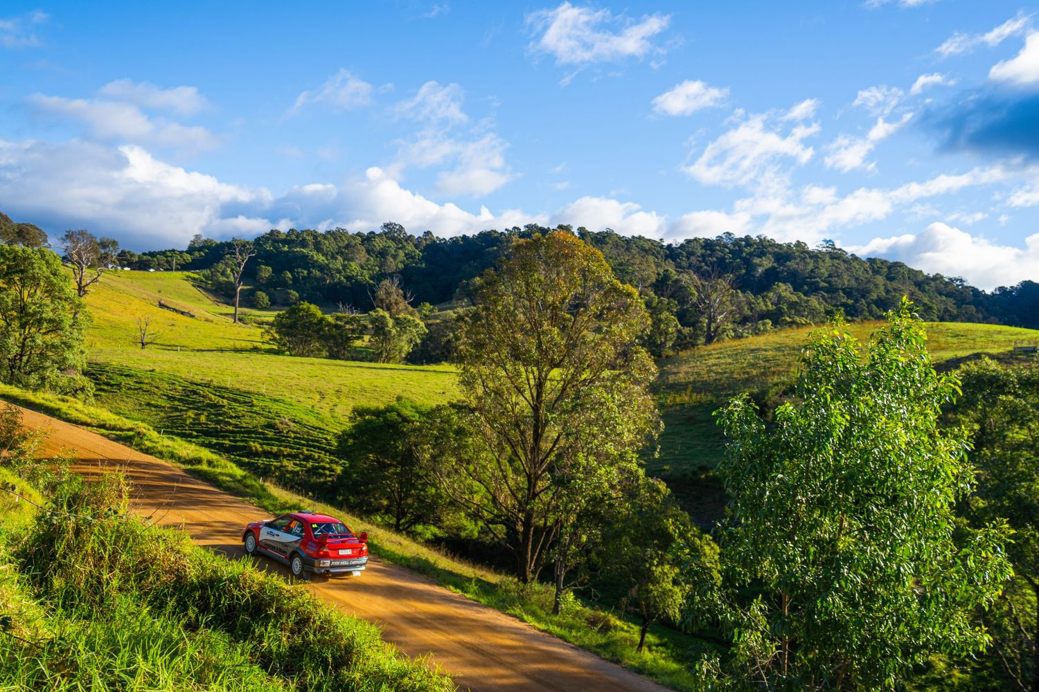 Red vehicle on dirt road winding through lush green hillside landscape.